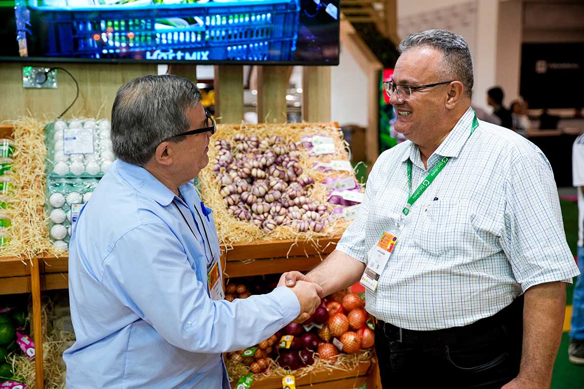 Two men are shaking hands and smiling in front of a market-style display of fresh produce, which includes onions, garlic, and eggs. One man is wearing a light blue shirt and dark glasses, while the other is in a white checkered shirt with a green lanyard and ID badge.