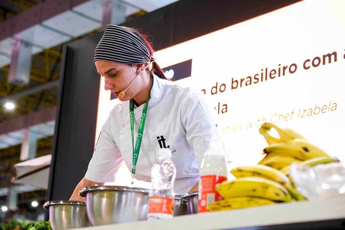 A chef wearing a white coat and a striped headband is preparing food at a demonstration table. A microphone is attached to the chef, and various ingredients, bowls, and a bunch of bananas are on the table. A large screen with text is visible in the background.