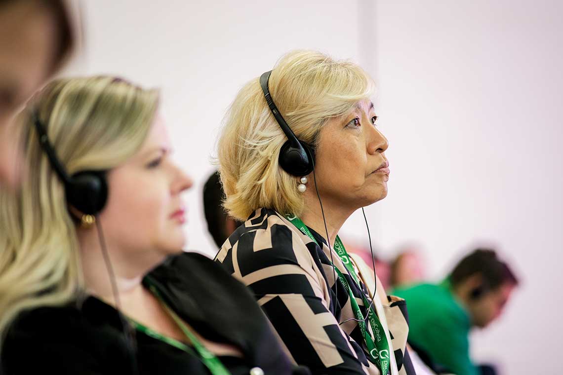 Two women are seated, facing right, wearing black headphones, and attending a conference. The woman in the foreground has blonde hair and is focused intently, while the woman behind her also listens attentively. Both have conference lanyards around their necks.