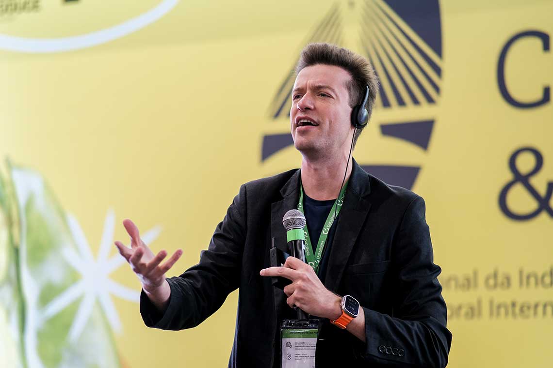 A person wearing a black blazer and a headset is speaking into a microphone at an event. They have a conference badge around their neck and are gesturing with one hand. The background features a yellow banner with text and graphics.