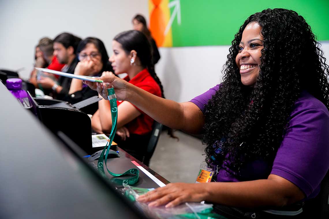 A smiling woman with curly hair wearing a purple shirt hands a lanyard to a person across a counter. Several other people are sitting at the counter in the background, engaged in various activities. The setting appears to be an event registration desk.