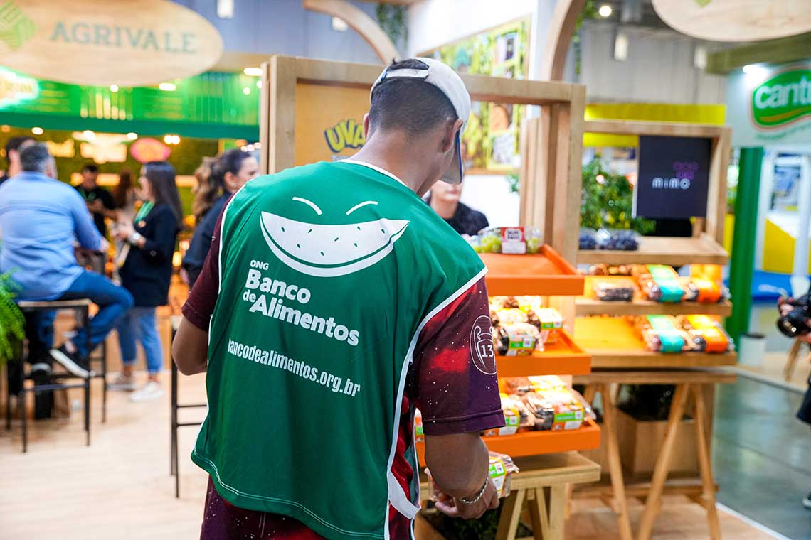 A person wearing a green vest with the logo and text "Banco de Alimentos" is standing in front of a display with various packaged food items. In the background, there are several people and booths at an indoor event.