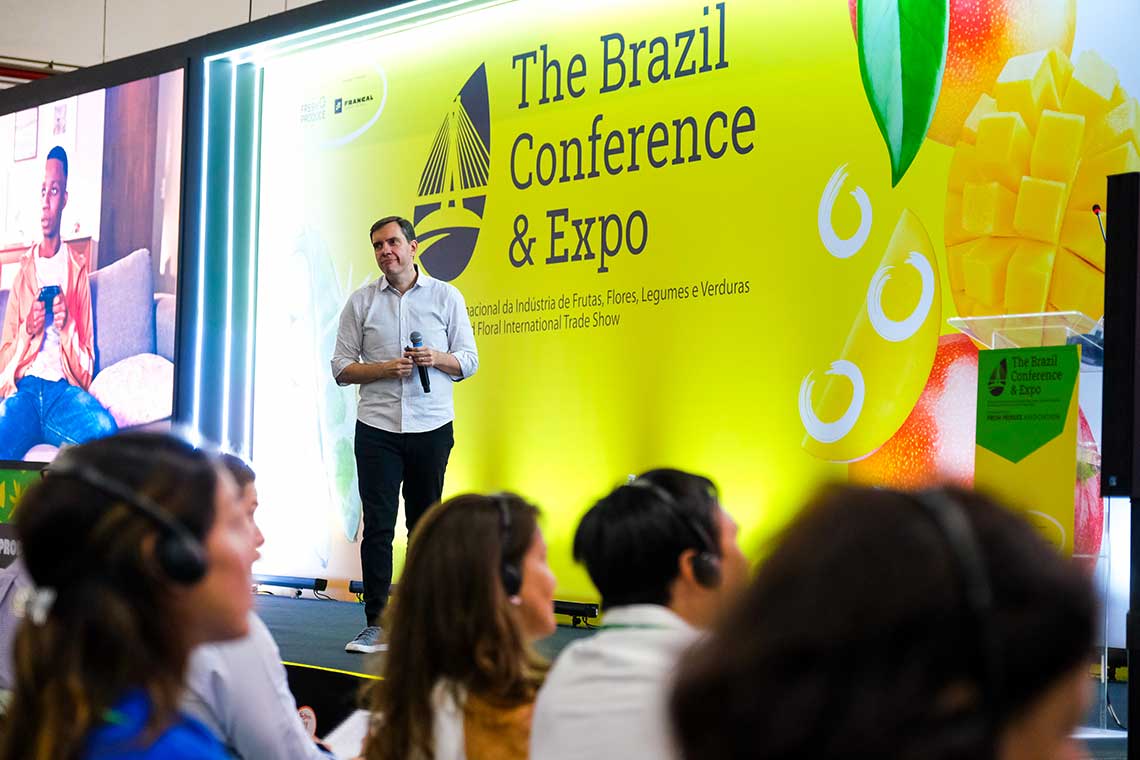 A man standing on a stage giving a presentation at The Brazil Conference & Expo. The backdrop is yellow with green accents and text. Attendees seated in the foreground are watching. Various fruits are displayed on the background banner.