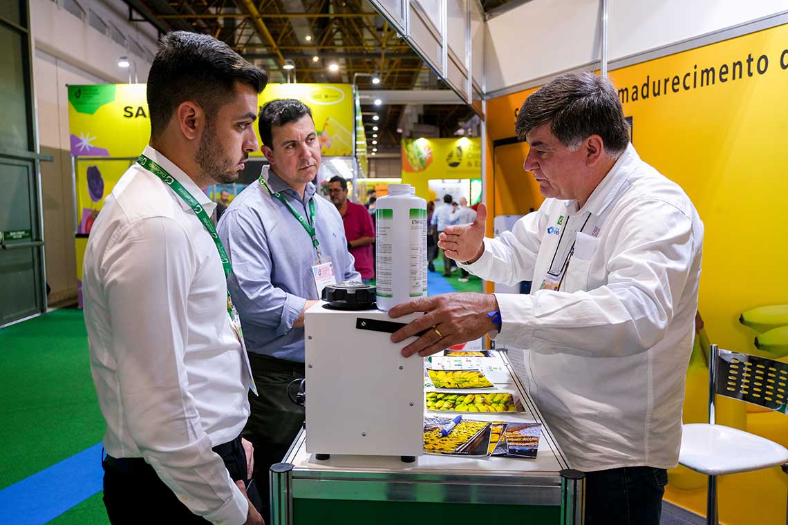 Three men are engaged in discussion at a trade show booth that displays agricultural products and equipment. One man, gesturing towards a white device with a bottle on top, appears to be explaining its features. The background shows colorful, agricultural-themed banners.