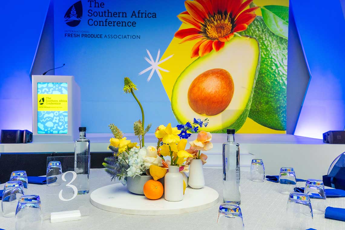 Conference table with flower and fruit centerpiece, against a backdrop featuring an avocado and flower image. Podium displaying "The Southern Africa Conference" text.