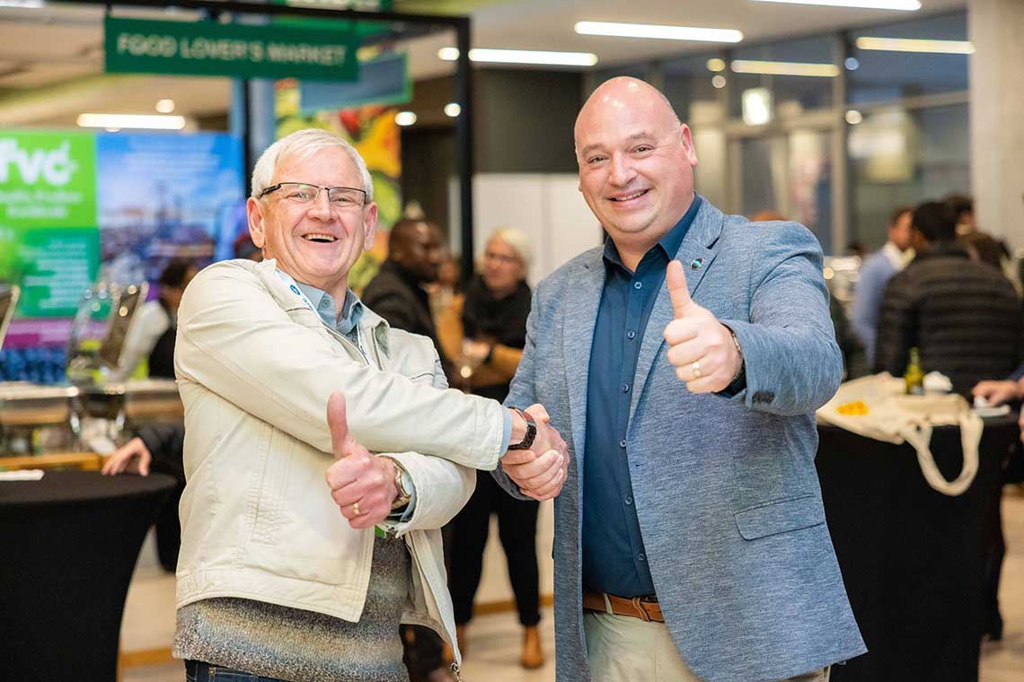 Two men at a conference give a thumbs up, standing in front of a sign that reads "FOOD LOVER'S MARKET."