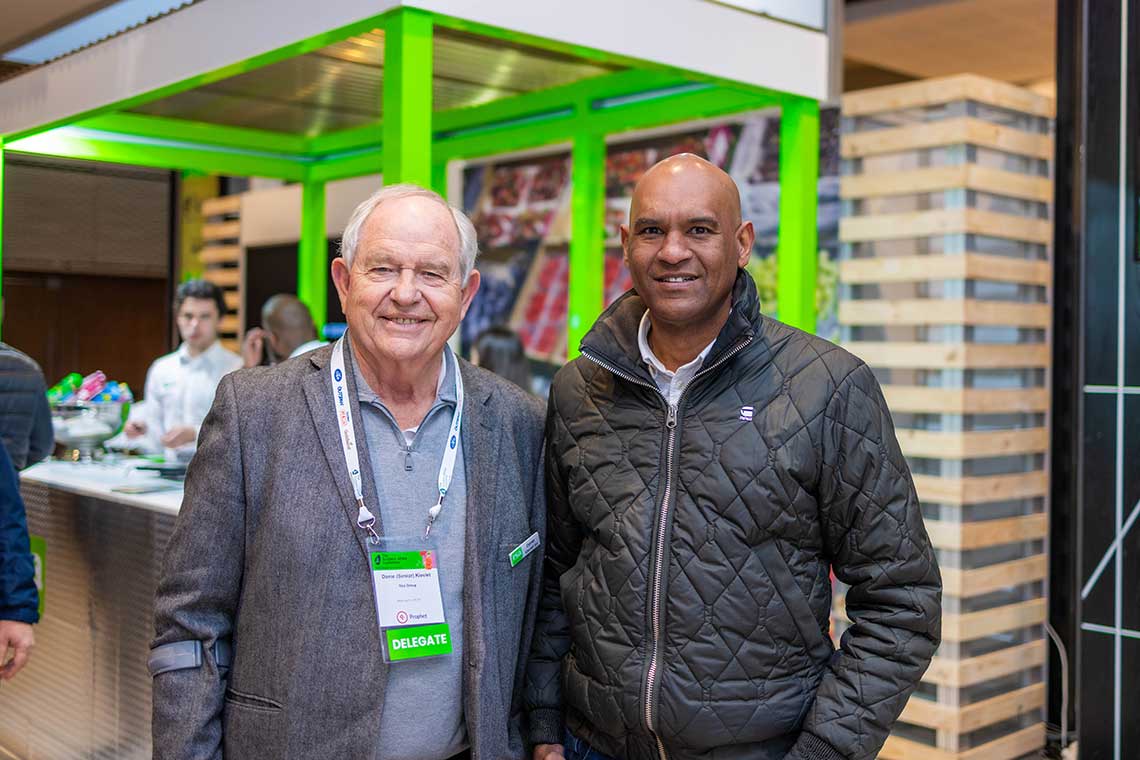 Two men smiling at a conference with a booth in the background.