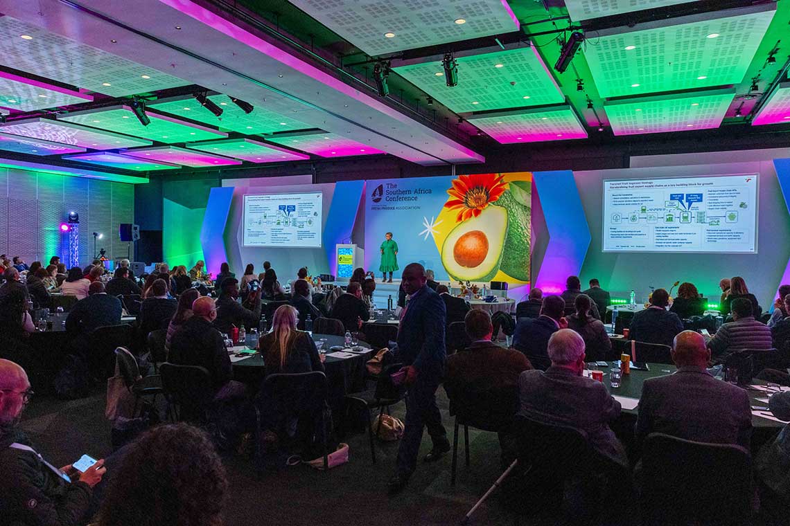 A conference room with a presenter on stage and attendees seated at tables, with colorful lighting and presentation screens.