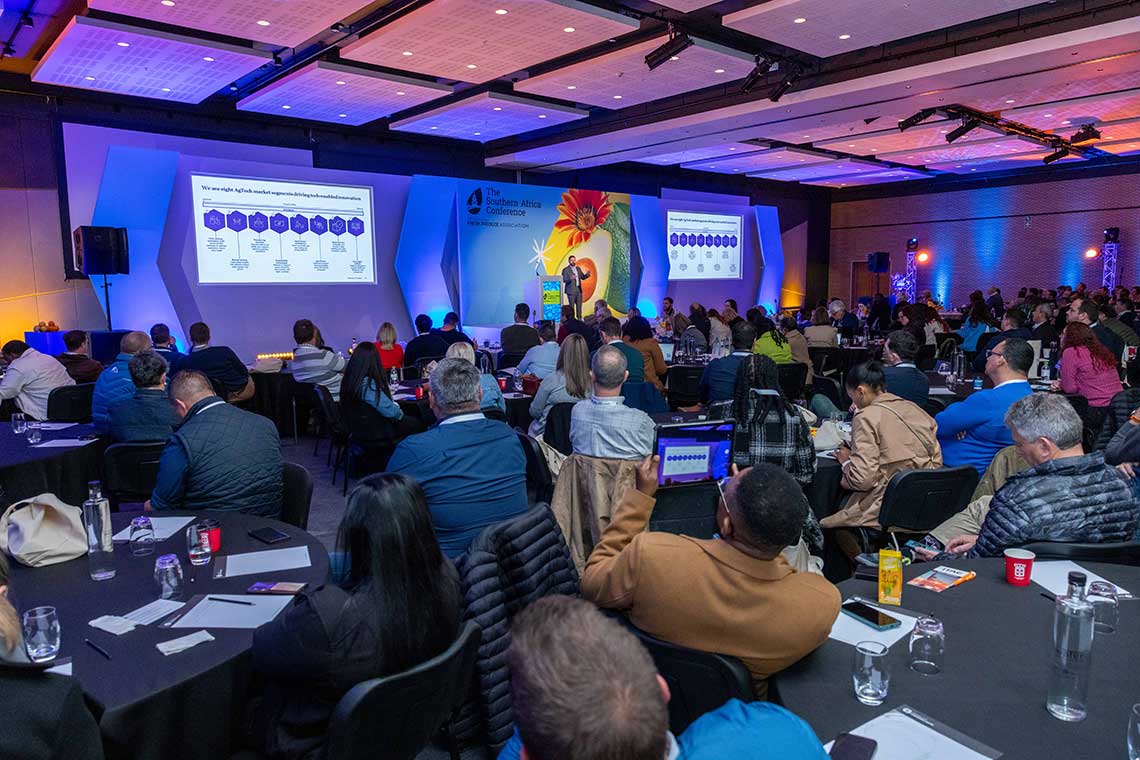 A conference room with attendees seated at round tables, watching a presentation on stage with screens displaying slides.