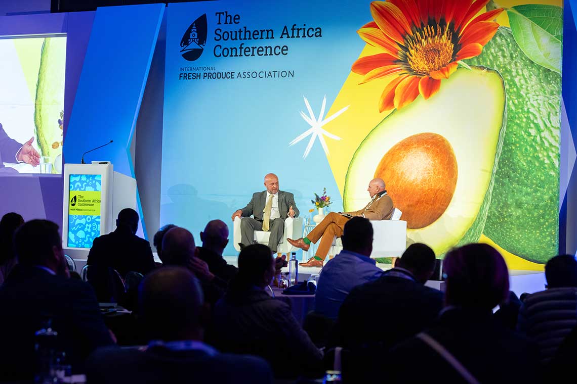 Two speakers sit on stage at a produce conference with an avocado-themed backdrop.