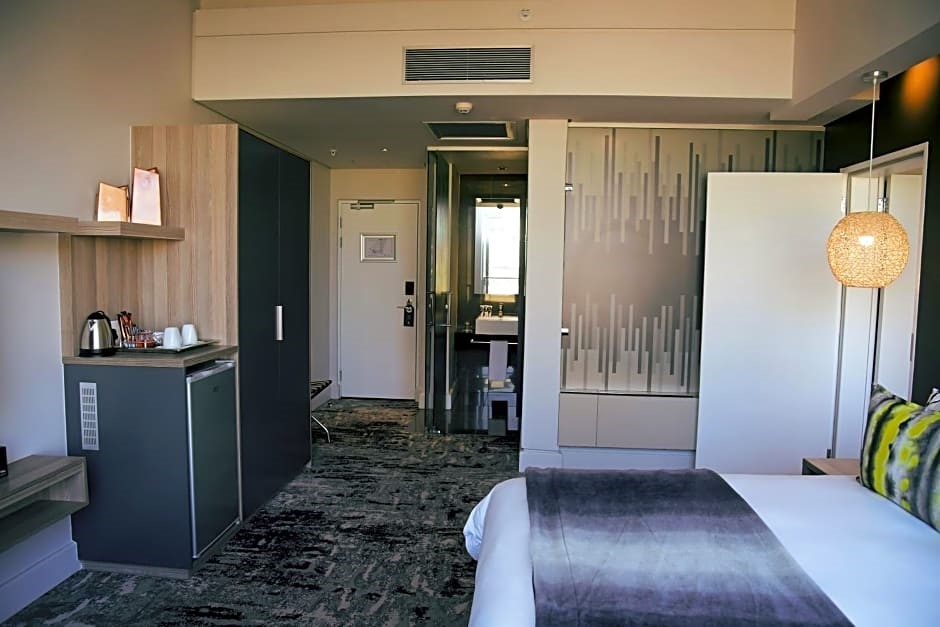 Modern hotel room with a bed, decorative pillows, abstract artwork, bedside lamp, and visible bathroom area.