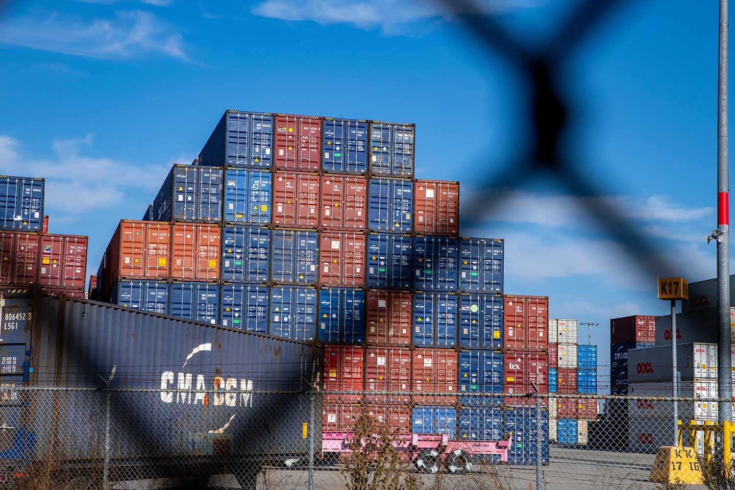 A stack of red, blue, and white shipping containers in a yard, seen behind a blurry chain-link fence, under a blue sky.